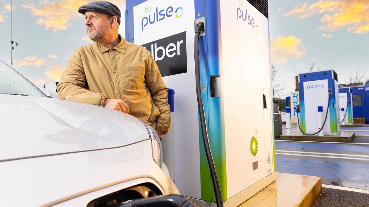 A man in a yellow jacket wearing a gray cabbie hat stands behind a white car plugged into an electric vehicle charger with a sign reading "bp pulse - Uber."