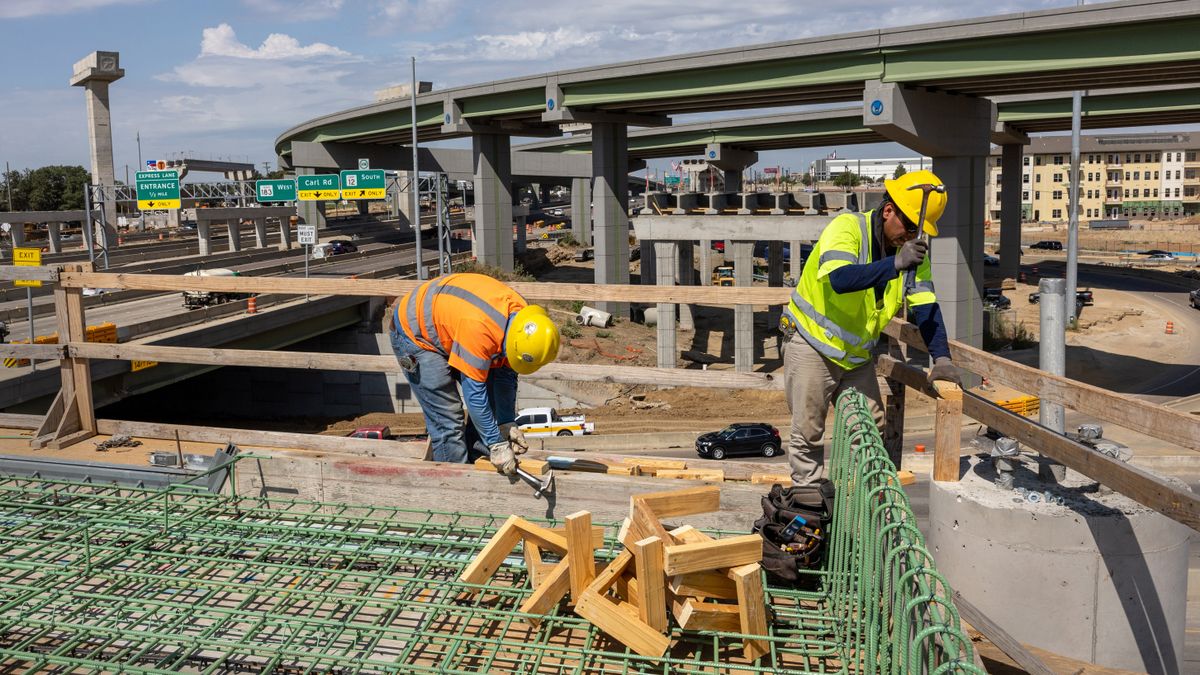 Two men wearing hardhats and bright vests are working on construction of a highway overpass in the foreground, while highways are seen behind them.